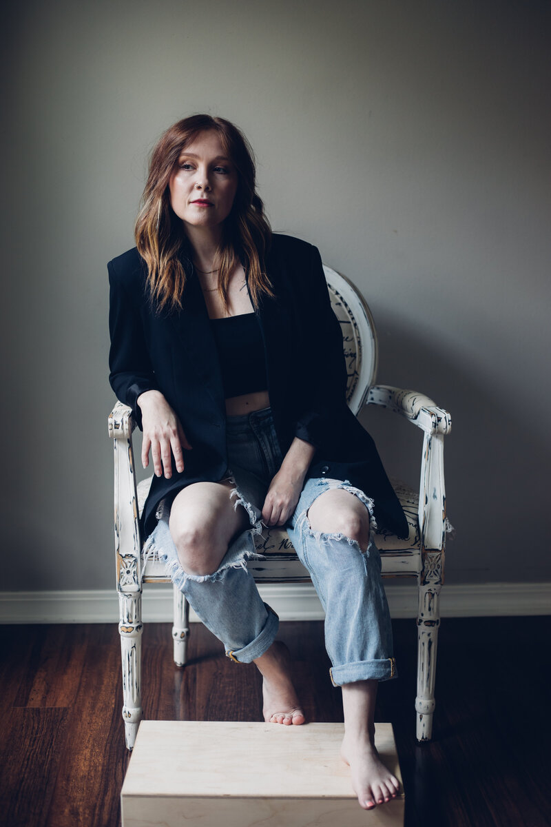 A woman with long hair, wearing a black blazer and ripped jeans, sits on an antique chair with one foot on a small wooden box, in a room with wooden flooring of an agency offering branding and web design for female entrepreneurs.