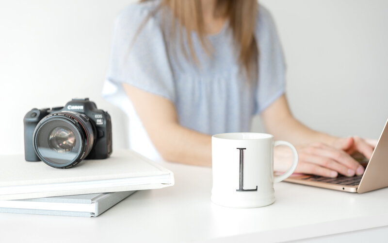 Liz is sitting at a desk working while a camera and a mug are in focus in front of her