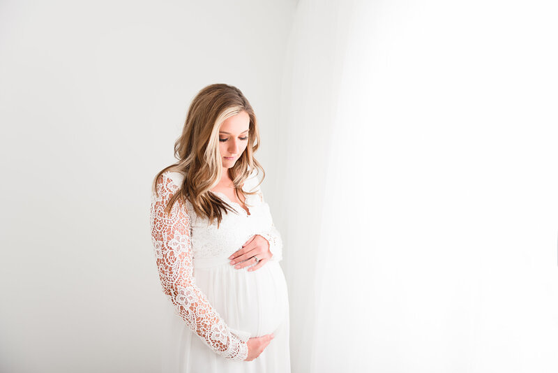 Pregnant woman in a white lace dress stands in soft natural light, gently cradling her belly. She gazes down, surrounded by a serene, minimalistic background.