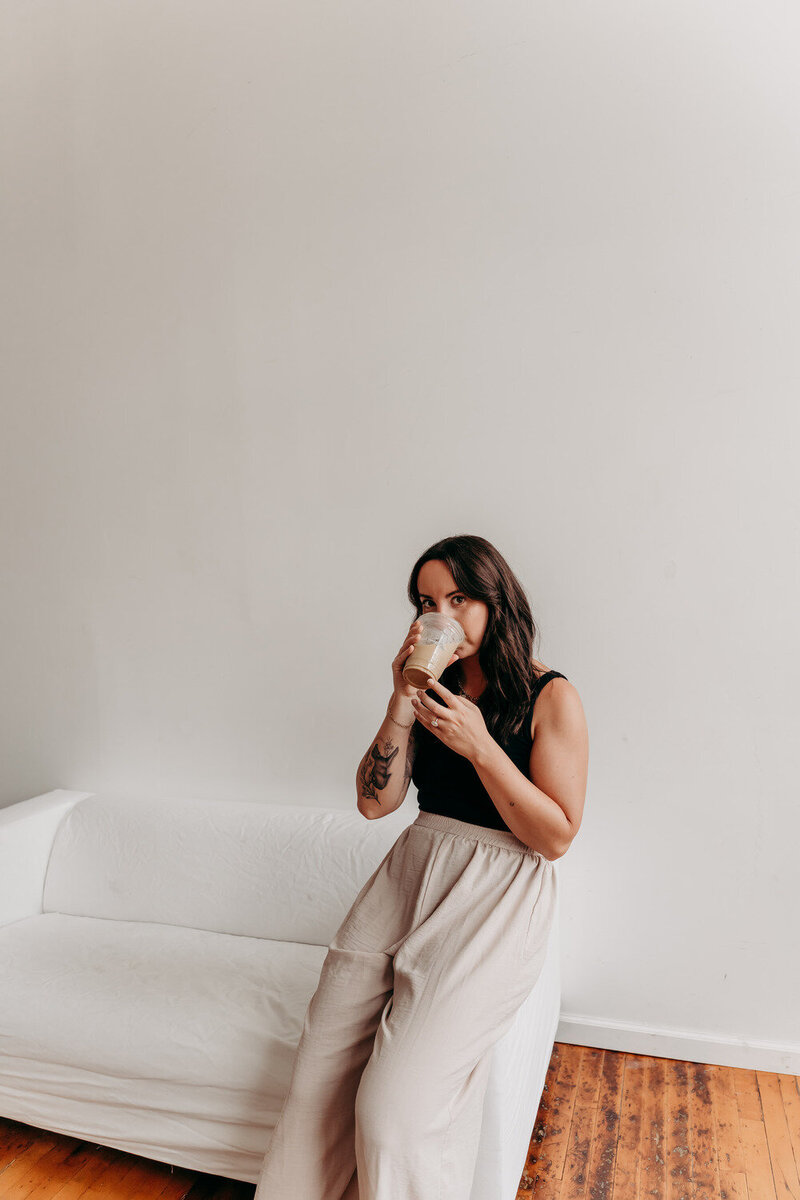 Delia, brand strategist, leaning against a white couch in a light studio space, wearing beige wide leg pants, a black tank top, sipping on an iced coffee.