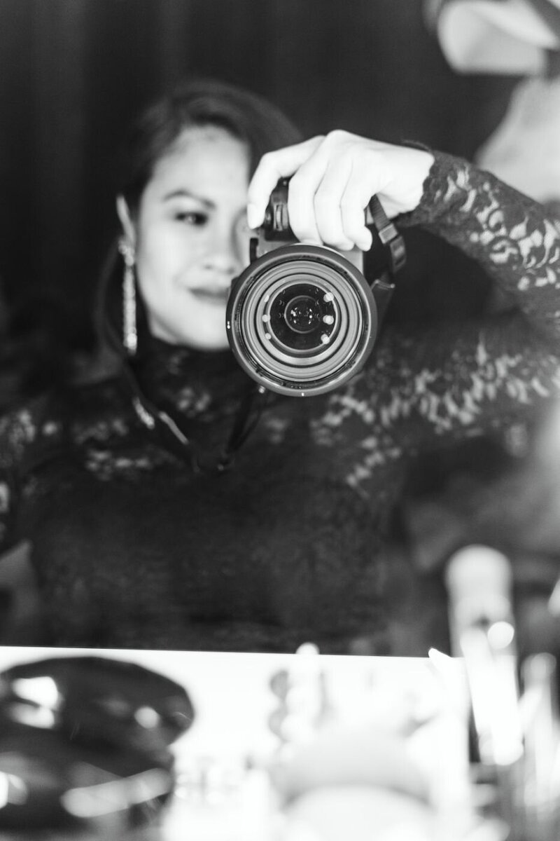 Black and white portrait of a female photographer in a black lace dress and gold earrings, holding a Sony camera in front of a mirror with makeup items on the table.