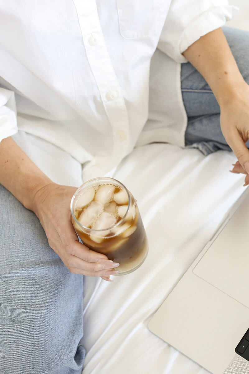 Woman sitting and holding a glass of iced coffee next to a laptop
