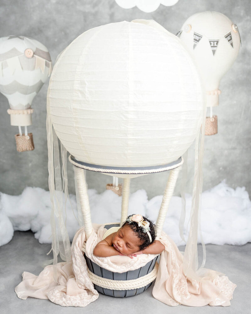 A peaceful newborn baby rests inside a basket designed as a hot air balloon, surrounded by soft, pastel-colored fabrics. The setup has clouds and additional balloon props.