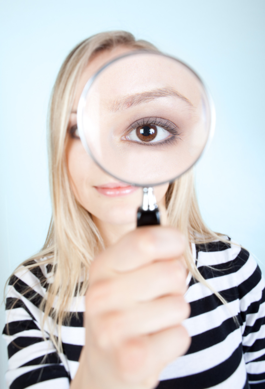 blonde woman wearing black and white striped shirt holding a magnifying glass in front of her face making one of her brown eyes look very large
