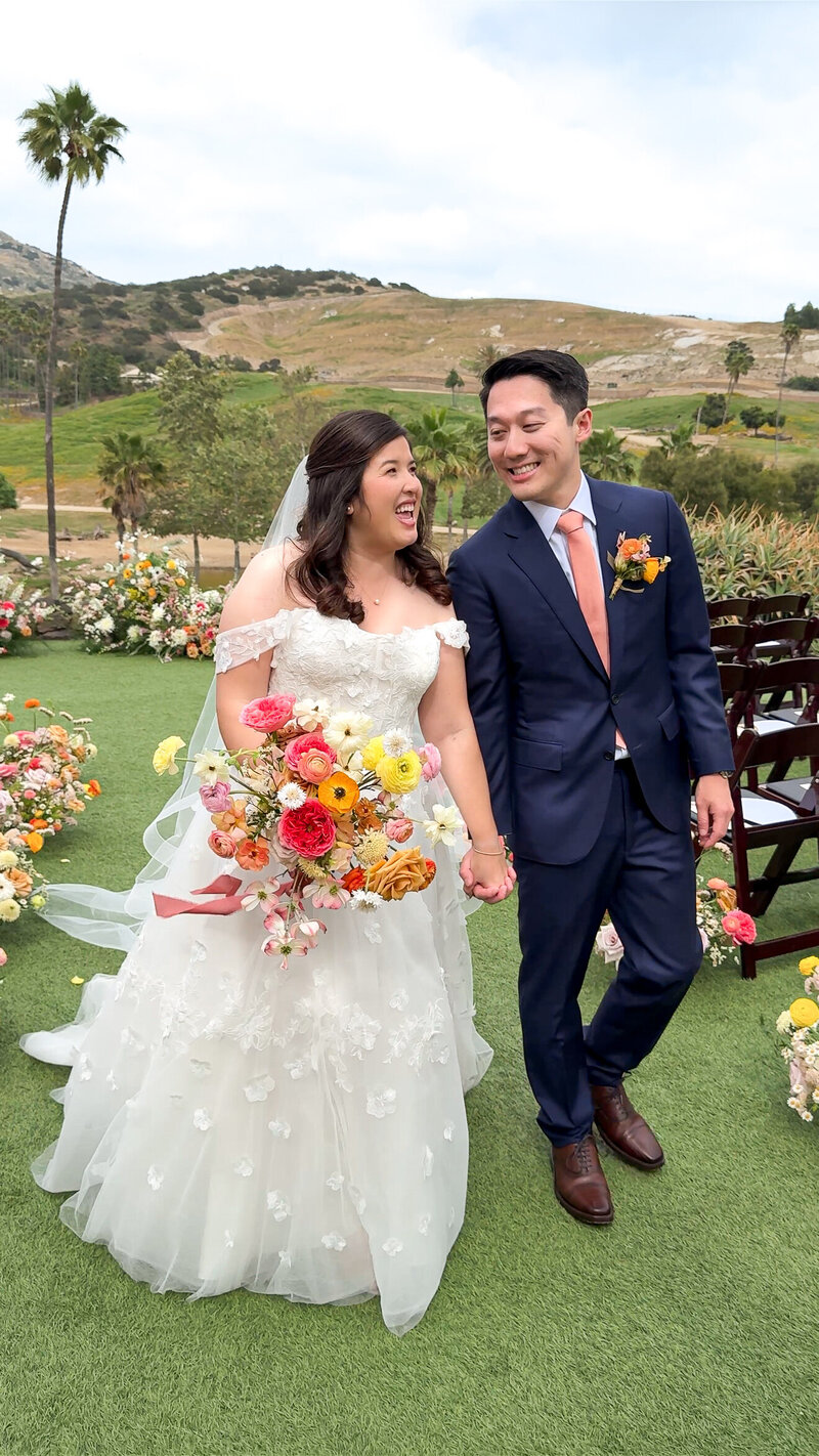 Bride and groom walking hand in hand by their ceremony set up holding floral bouquet at San Diego Safari Park in Escondido, California.