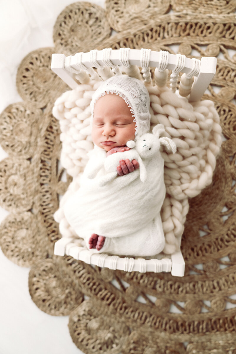 Newborn bliss: a serene baby sleeps soundly swaddled in white, cradled on a lacey background with a plush bunny by their side.