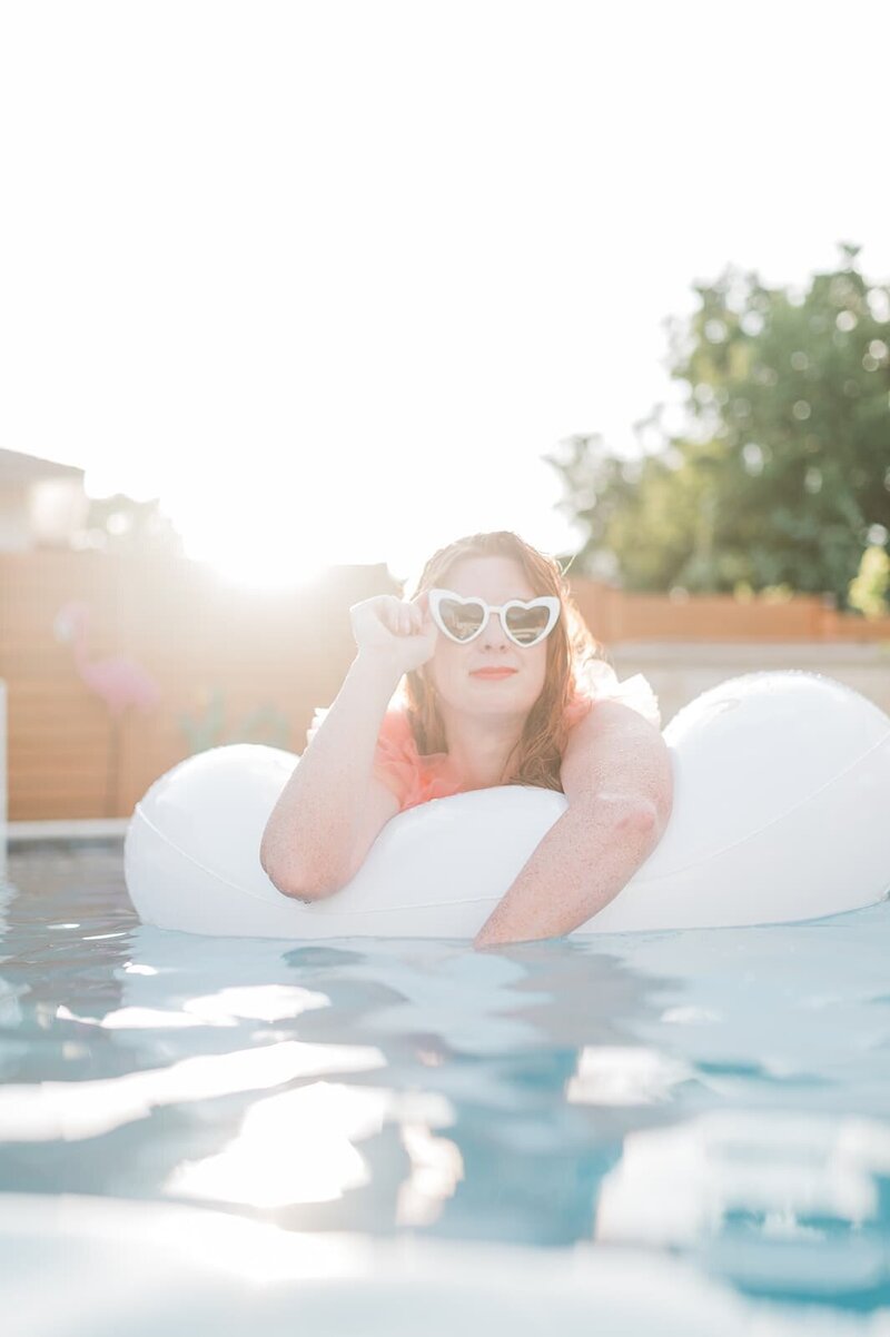 A woman with heart-shaped sunglasses relaxes on a white pool float, bathed in soft pink lighting, exuding a dreamy summer vibe.