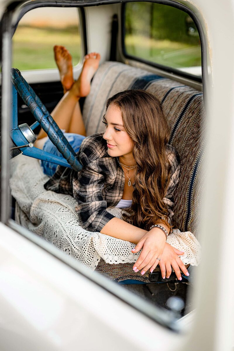graduate portrait of a senior girl looking off camera as she lays on her belly in a truck seat wearing a flannel and jean shorts