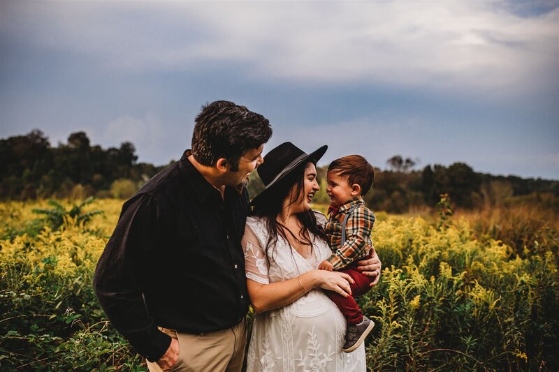 Baltimore photographers  captures family portraits in a lush summer field with mother holdin gher toddler and laughing with him as the father leans into them and smiles