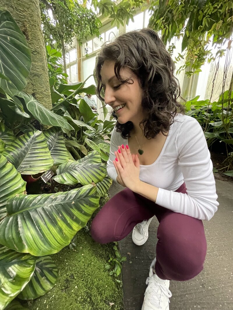 An intuitive healer stands in a beautiful botanical garden surrounded by lush greenery. She stands with her hands in prayer position, gazing down at the large leaves above her with a serene smile on her face. The natural setting and the healer's peaceful expression create a sense of calm and tranquility.  [Nicole QW]