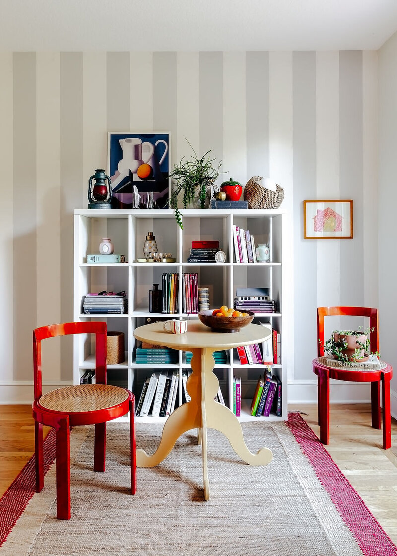 A shelf with colorful books and knickknacks in front of a striped wall with a small dining table in the forefront, representing the colorful home decor found in Erin's home. 