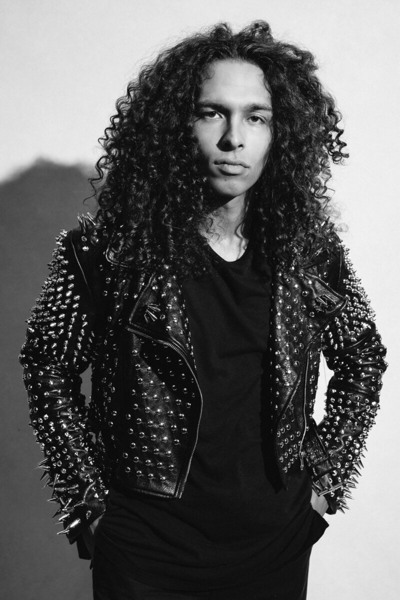 Black and White image of man with long curly hair with a serious look on his face. Wearing a black t-shirt and a black leather jacket with spikes all over it. 
