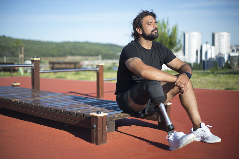 This image shows a man with a prosthetic leg, wearing dark athletic clothes and white tennis shoes, sitting on outdoor exercise equipment. His eyes are closed, and his face is turned up toward the sky. A blue sky and cityscape are visible in the background.
