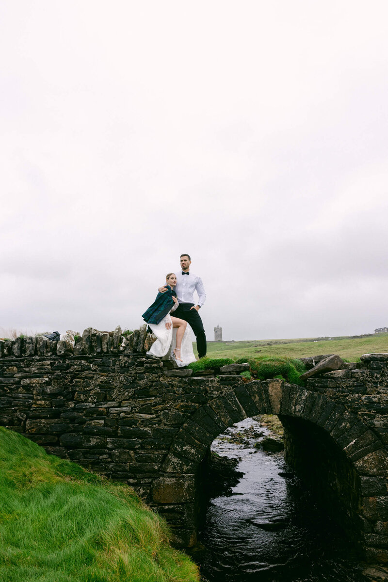 A couple posing for a photo during their elopement at the cliffs of moher the bride is sitting on a stone wall with her grooms jacket covering their shoulders
