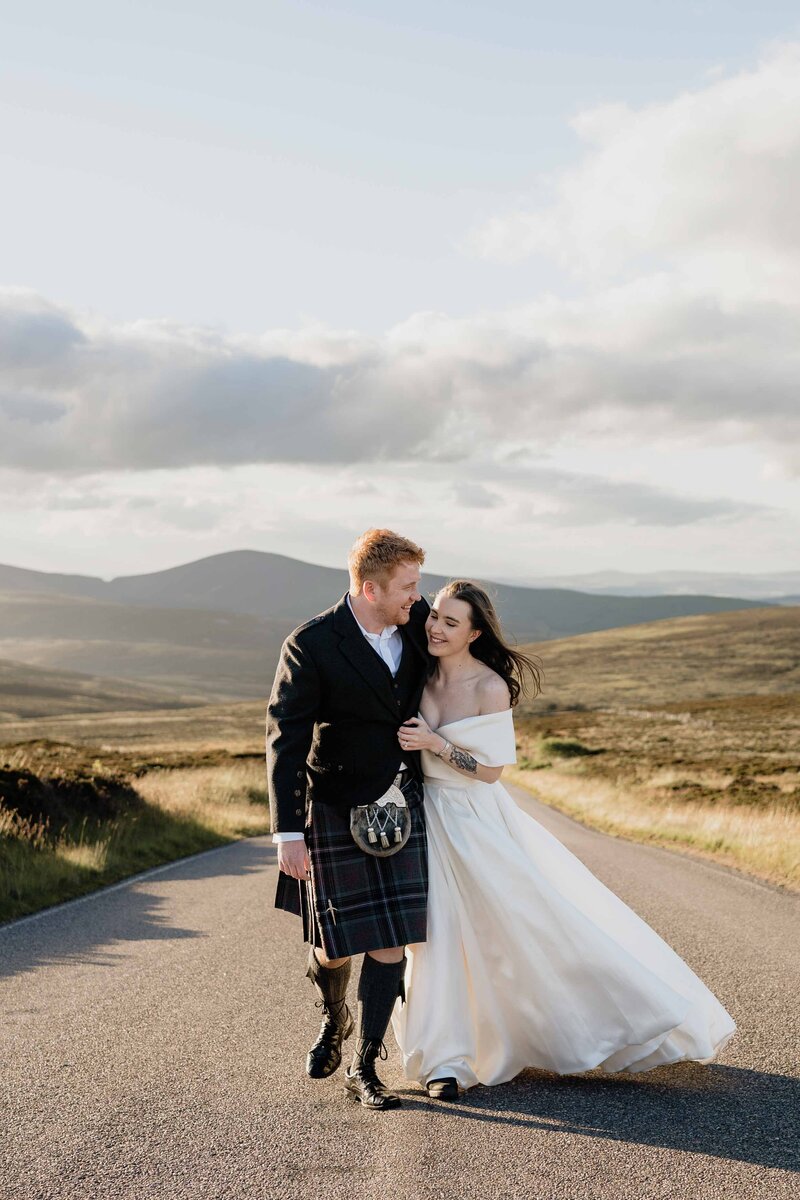 A newly married couple walk along a mountainous Aberdeen road. Mountains can be seen behind them as they smile towards their wedding photographer.BTQ+ couple walk down stairs hand in hand. They pose for wedding photographs in a city centre garden.