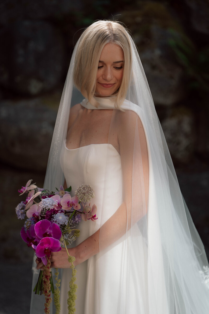 Bride in modern cape veil and strapless gown holding vibrant bouquet with orchids and wildflowers