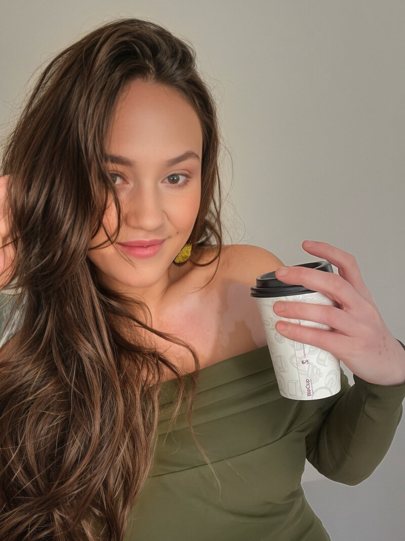 a brunette woman looking at the camera holding a coffee in her hand smiling, wearing a green off shoulder dress