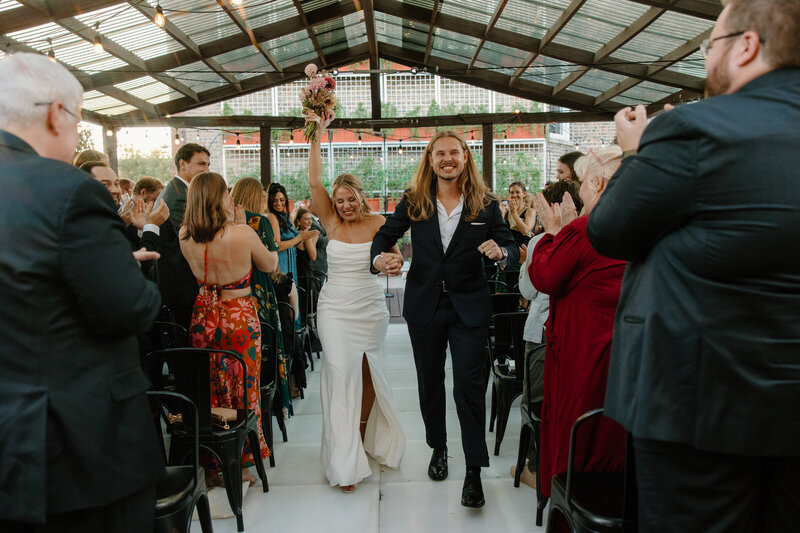 Bride in strapless satin wedding dress with a high slit raises her bouquet above her head in celebration as she walks down the aisle with her new groom and the guests applaud.