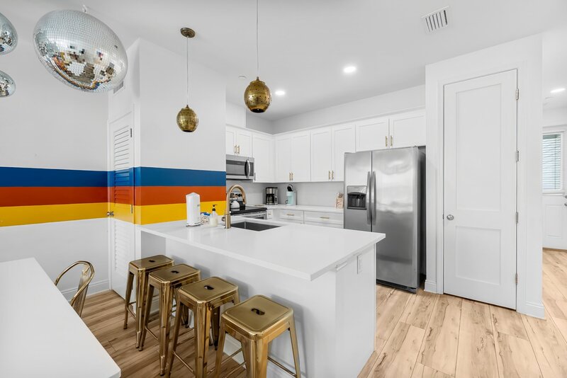 Full kitchen with white cabinets, stainless steel appliances, and a rainbow-striped accent wall with gold pendant lights and stools.