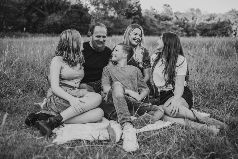 A family of five is sitting on the ground, talking and smiling, getting family photos done in a field in OKC