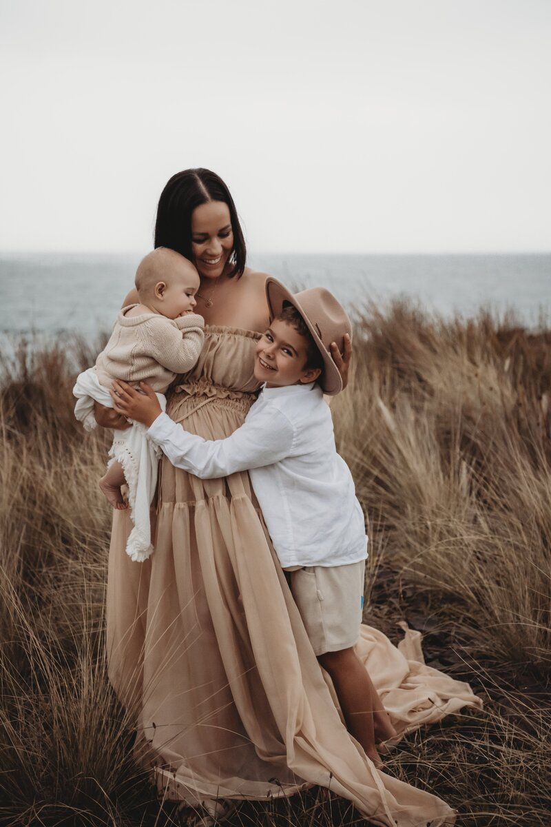 A woman holding a baby and embracing a child in a hat, standing in grassy dunes by the sea, captured beautifully by a Wollongong motherhood photographer.
