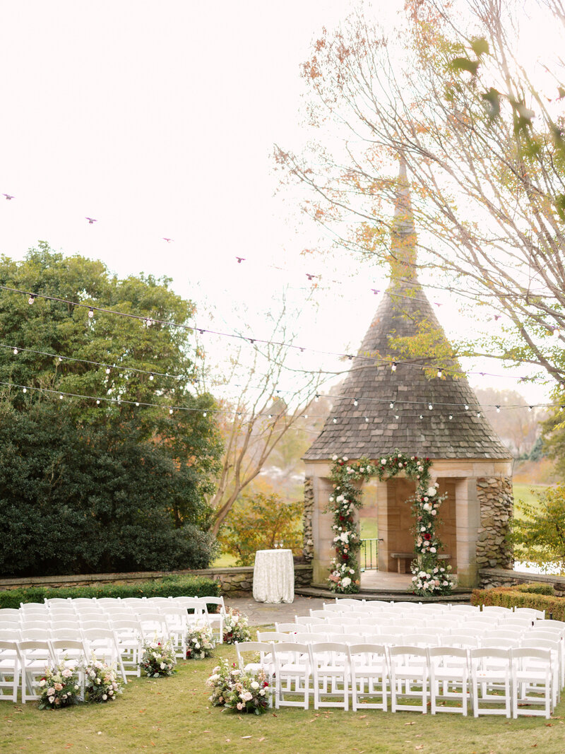 November wedding at Graylyn Estate, designed and planned by Charming Carolina — full ceremony view of the White Garden with gazebo altar florals, ground arrangements down the aisle, string lights overhead, and white garden chairs set for guests.