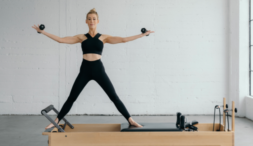 Andrea standing wide-legged on a reformer with a weight in each hand