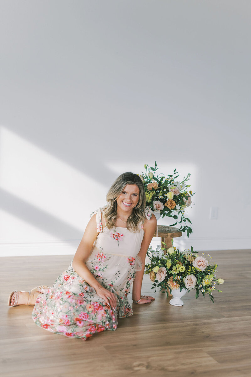 Headshot of Lindsey Kristian in a floral dress sitting on the floor next to colorful flower arrangements
