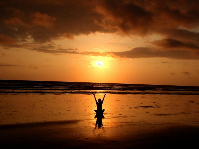 Silhouette of a woman with her arms in the air while sitting on the beach looking at the water.