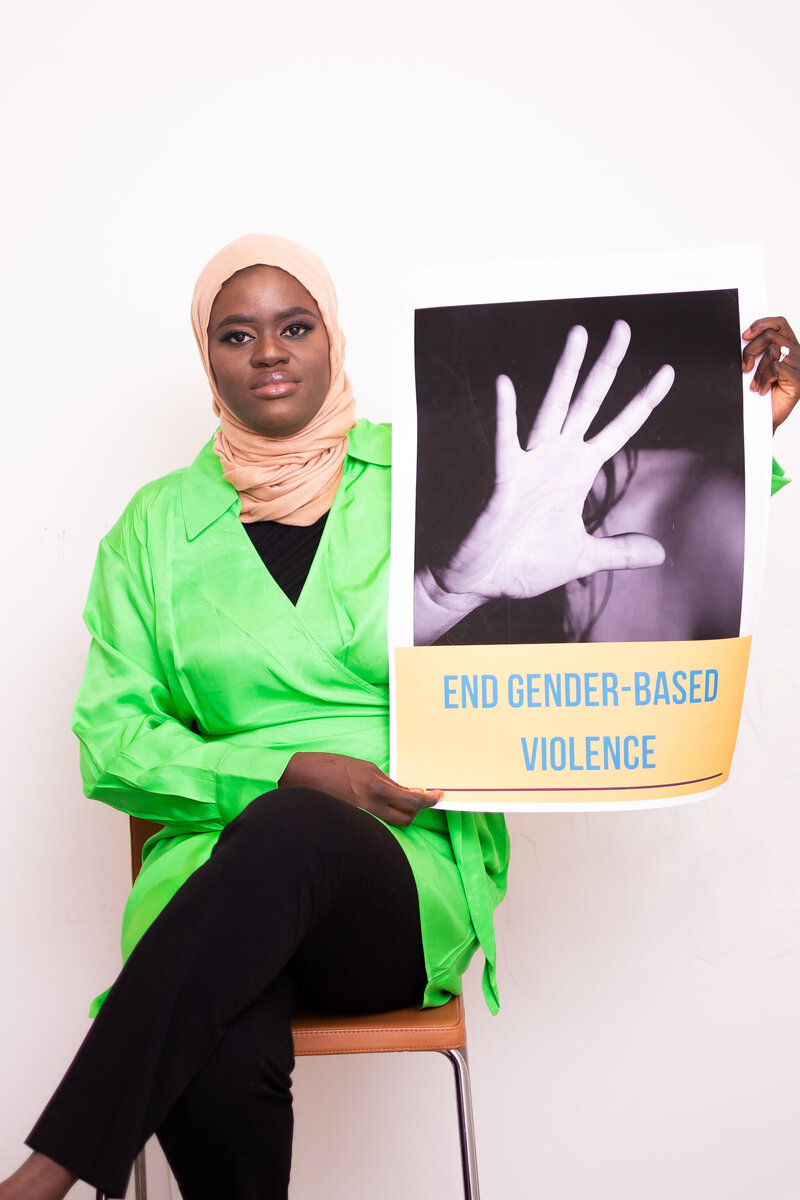 Picture of Isatou Bittaye Jobe, a grant writing consultant, sitting and wearing a lime green tunic. She is holding a sign that says "end gender bases violence".