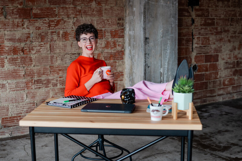 Wedding planner in bright orange top and pink pants with feet resting on desk
