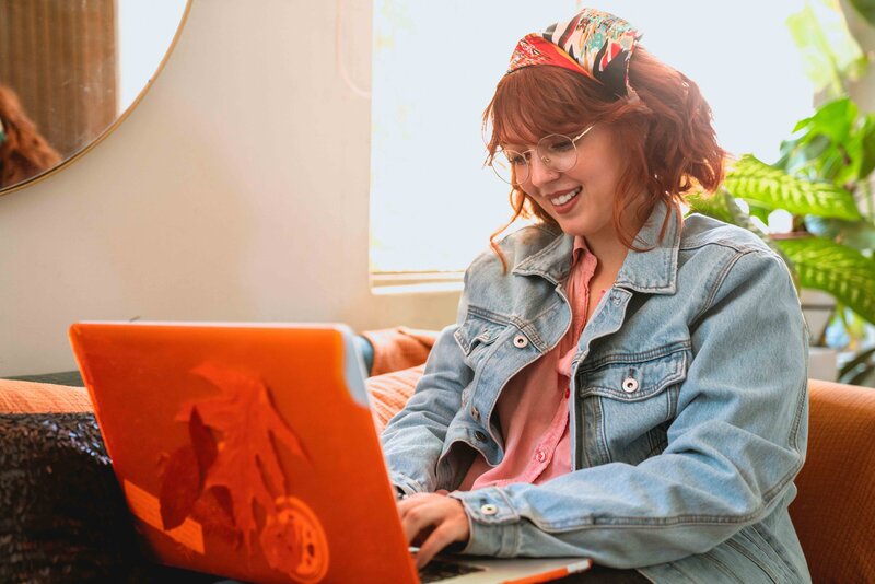 This image shows a young feminine-presenting white individual sitting on a couch, typing on a laptop and smiling.