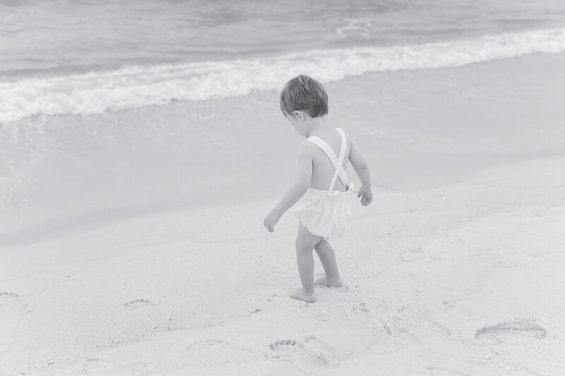 Timeless beach childhood photography—little boy in a vintage romper walks along the sandy shore, leaving footprints by the waves. Orange Beach, AL family photographer capturing light-filled, coastal moments