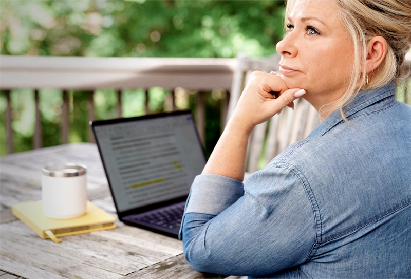 Chanda Bell sitting at a table with her elbow on the edge. She has her arm bent up and her chin resting on her hand. In front of her is an open laptop with text on the screen. Next to the laptop is a notebook with a mug sitting on top. Behind her is a deck railing and trees.