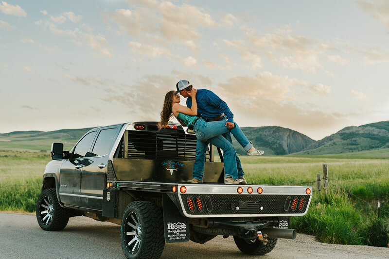 Eastern Idaho couples photographer Adriana Montoya Photo. A man is holding his girlfriend in a dip on the bed of their truck. The girl is smiling and has her hand around his neck. Behind them is a small mountain with greenery.