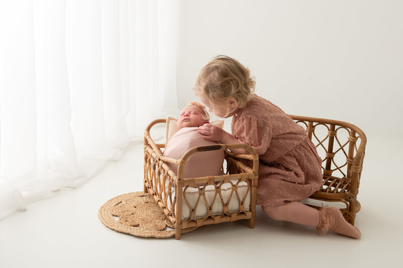 newborn baby in een rieten bedje met grote zus in een minimalistische witte setting gefotografeerd door Digidroom in Malle