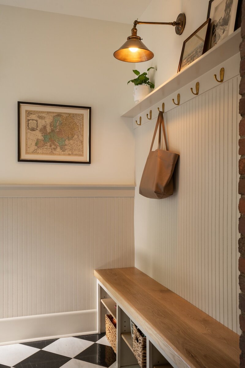 Mudroom with fluted white accent wall, golden hooks, wooden bench with storage, white and taupe half-tone walls, and decorative wall shelf.