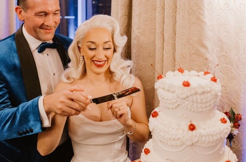 Bride wearing a vintage-inspired blonde wig smiles while cutting her wedding cake, styled with soft glam waves and classic elegance.