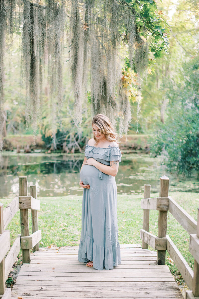 Woman in an off the shoulder light blue dress holding her pregnant belly in front of a pond