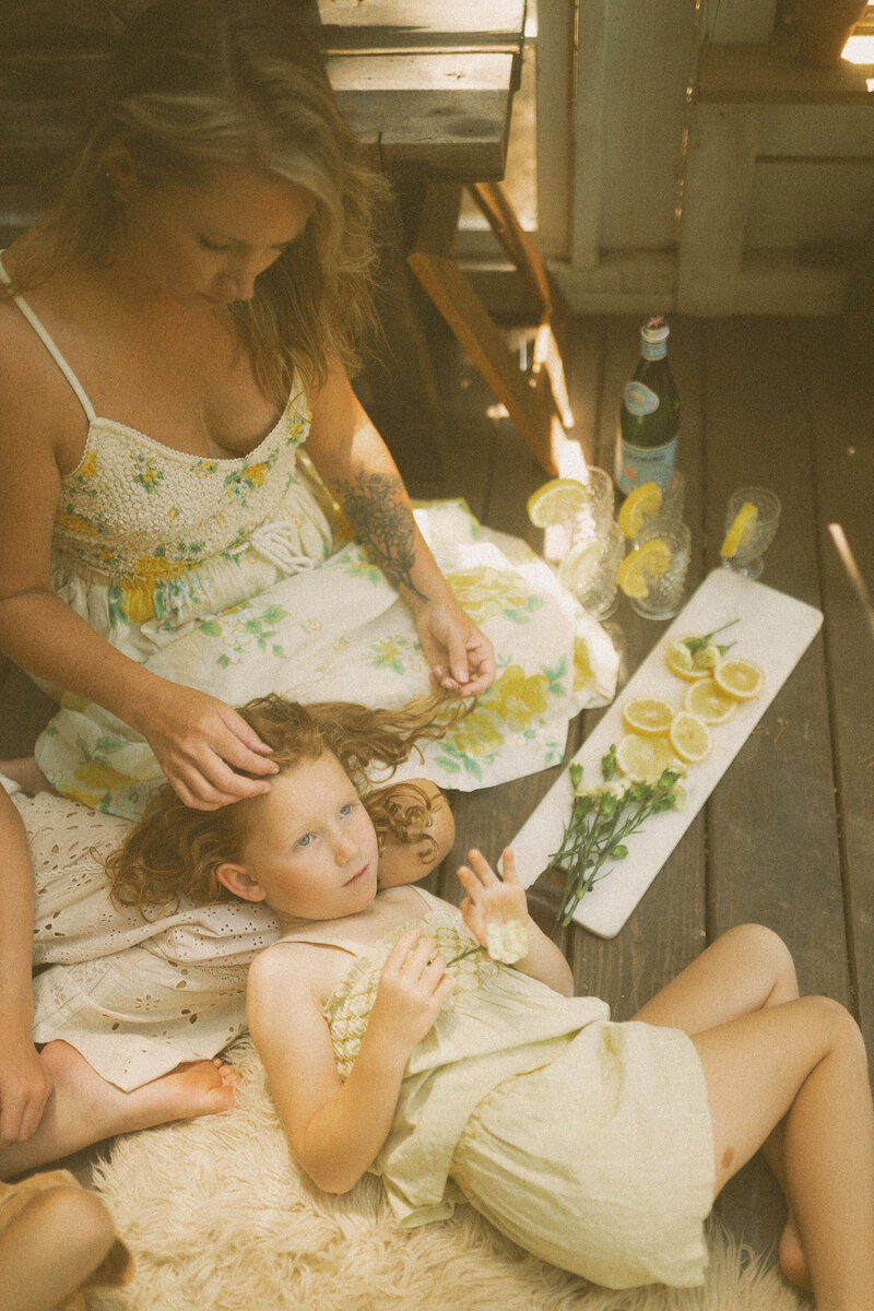 A girl laying her head in her mom's lap while her mother plays with her curly red hair. 
