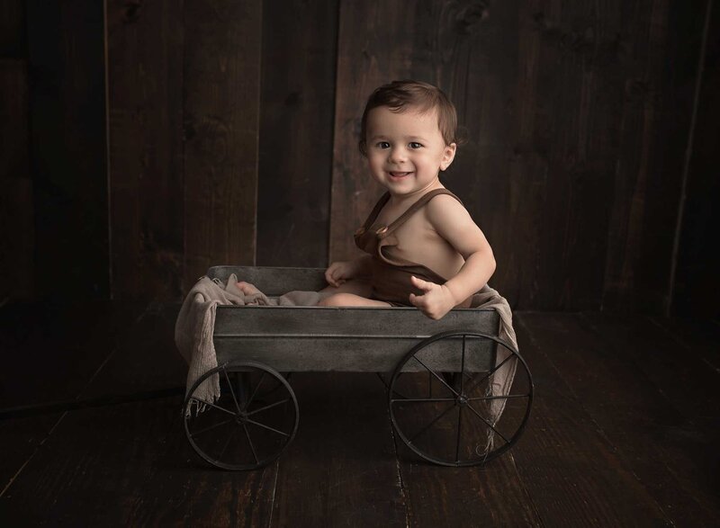 A young toddler boy in brown overalls sits in a small tin wagon in a Lafayette Baby Milestone Photographer studio