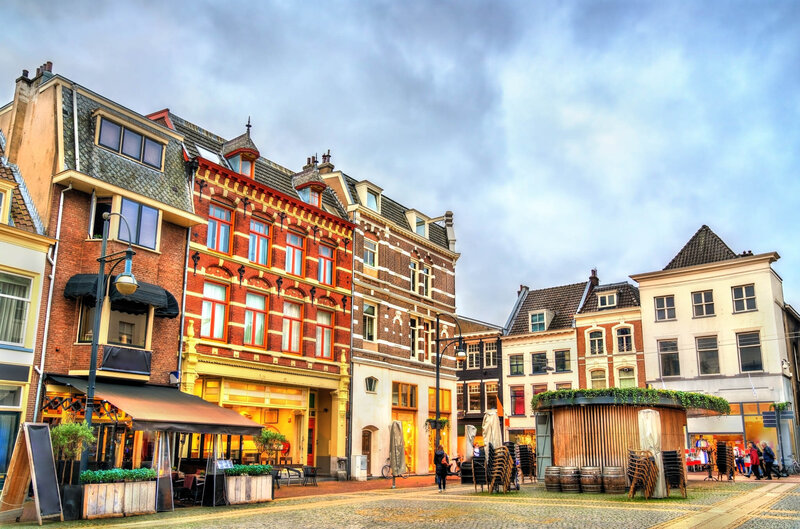A colorful row of historic European buildings surrounding a lively town square, with cafés, outdoor seating, and people walking under a cloudy sky.