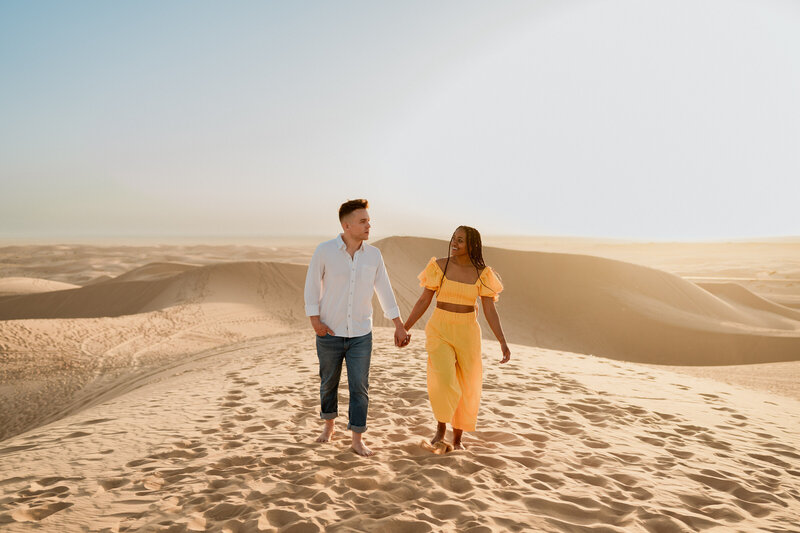 A couple holding hands and walking along sand dunes.