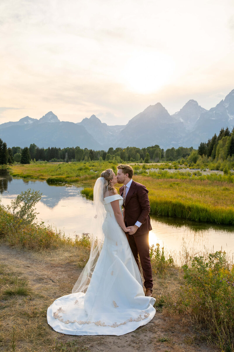 Elegant bride in an off-the-shoulder gown and groom in a tailored brown suit at sunset, with the tranquil waters reflecting the towering Grand Teton peaks in the background, creating a luxurious Jackson Hole wedding moment.