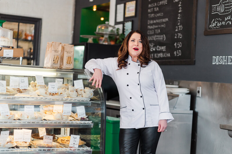 Candid image of a female chef leaning against a display of baked goods at her cafe