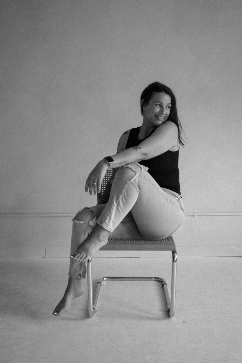 Brunette woman in jeans and black tank top with one foot up on the chair looking away