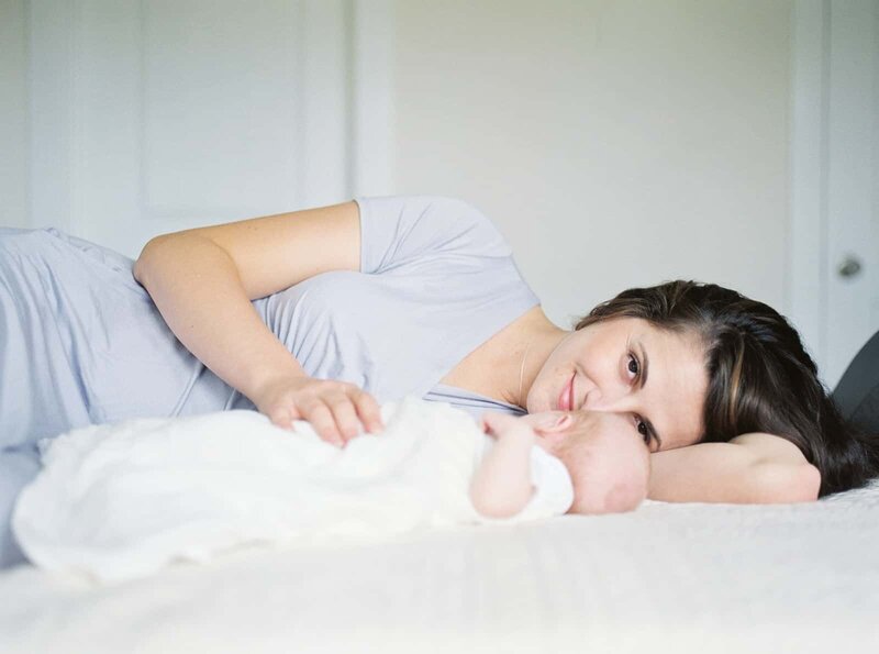 A young mother lays on her bed with one hand on her newborn and looks at the camera during her newborn session by DC Newborn Photographer Marie Elizabeth Photography