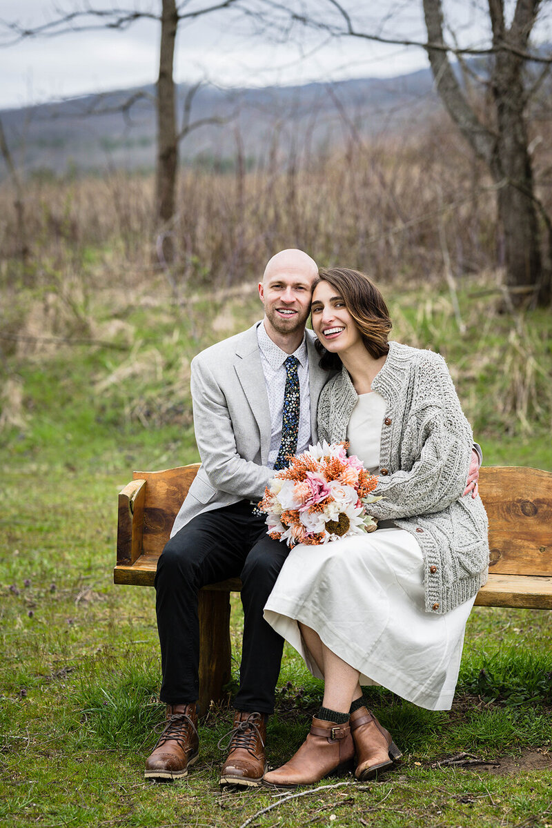 A couple on their elopement day in Virginia at Heritage Park sit on a nice wooden bench and smile for the camera.