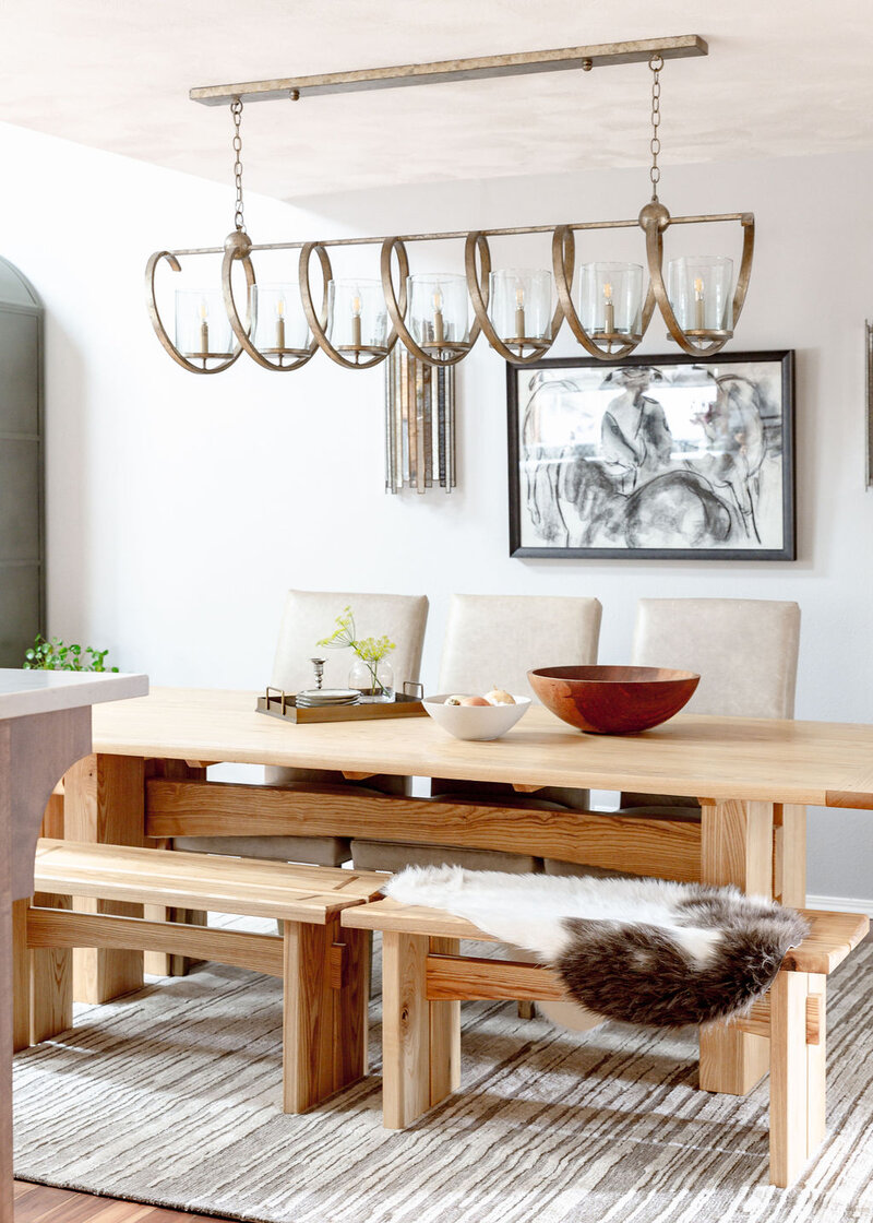 A dining room featuring a wood trestle table with benches, a hanging light fixture, and woven run representing a nordic interior design by Winsome Home Interiors.