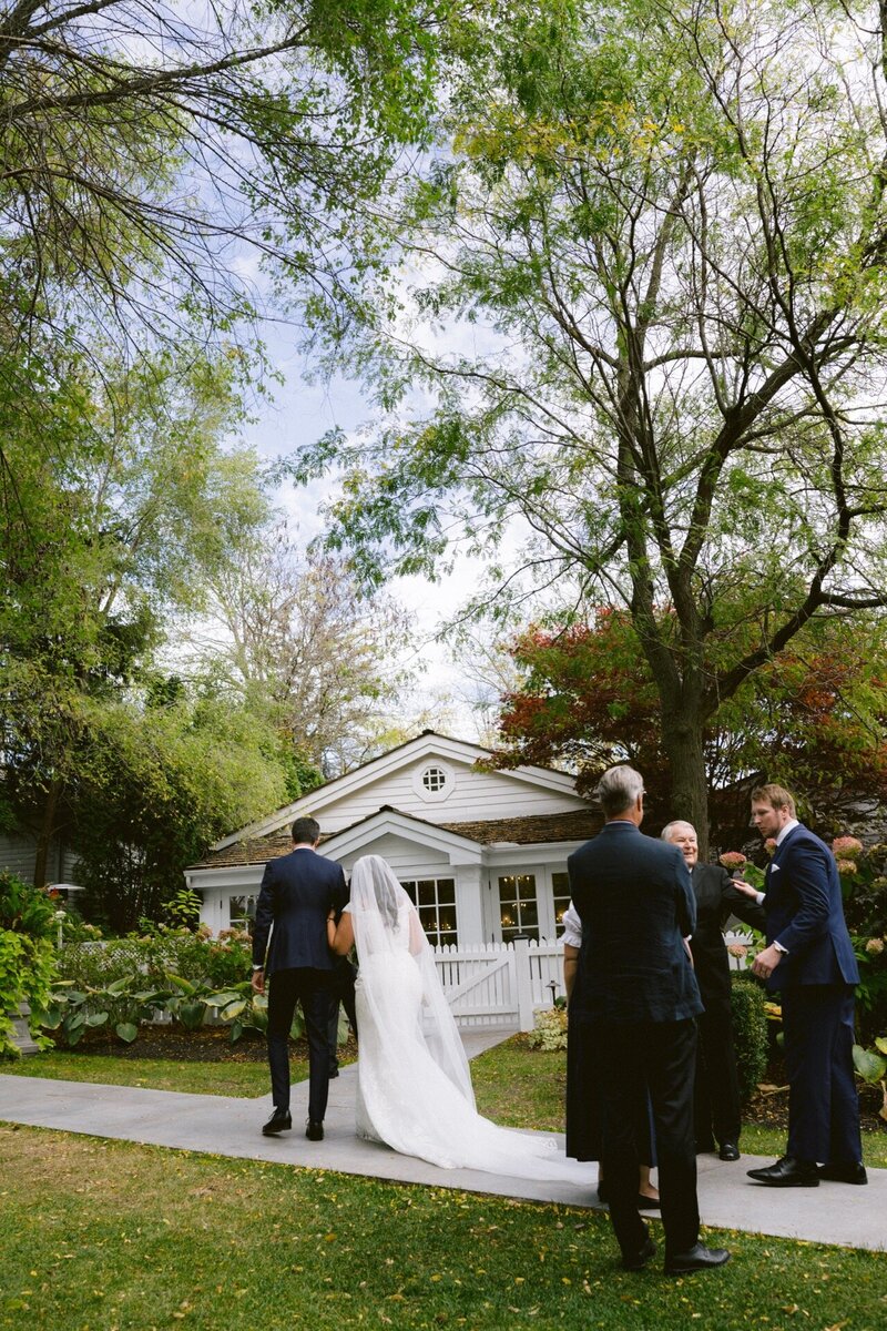 A bride walking towards a wedding ceremony outdoors with guests looking on.